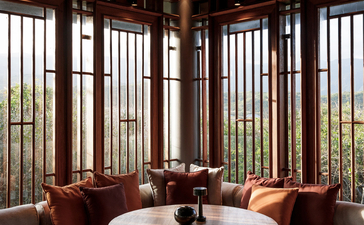 Amanoi gallery space with round table, wooden chairs, and floor-to-ceiling windows overlooking landscape.