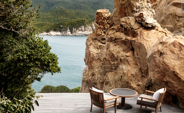 Two wooden loungers on a wooden deck beside a dramatic rocky outcrop, with calm turquoise waters and forested hills beyond at Amanoi.