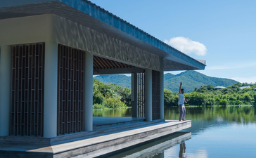 A modern pavilion with dark timber and blue-tiled roof extends over still waters at Amanoi, reflecting clear skies and distant hills.