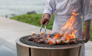 Person grilling fresh catch over an open flame at Amanoi, with coastal views beyond.