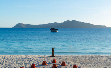 安缦璞乐度假村海滩上，客人们围坐在沙滩，面向平静的蓝色海水和远处的山峦。