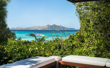 安缦璞乐度假村的平台眺望清澈蔚蓝的海湾，棕榈树和绿植环绕，木制栏杆在前。