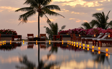 Amanpuri's waterfront at dusk, with palm trees reflected in still water and warm lights illuminating the resort buildings.