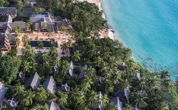 Aerial view of Amanpuri resort nestled along a turquoise coastline with sandy beach and forested grounds.