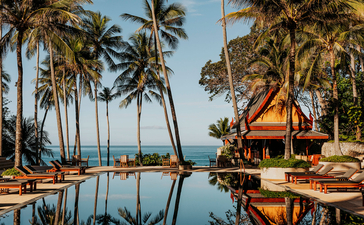 Amanpuri's infinity pool stretches toward the Andaman Sea, framed by tall palms and teak pavilions in golden afternoon light.