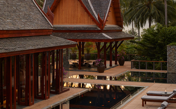 Amanpuri dining pavilion at sunset, with table settings arranged on a wooden deck beneath traditional Thai architecture and palm trees.