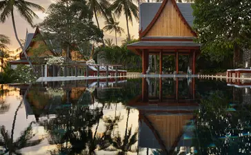 Thai sala reflected in still water at Amanpuri resort, surrounded by palm trees at dusk.