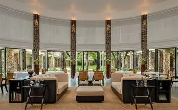 Amansara's dining venue with floor-to-ceiling windows overlooking gardens, featuring dark wooden pillars and contemporary furnishings.