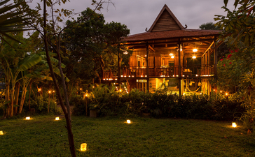 Amansara's dining venue illuminated at dusk, with warm golden lights glowing across the colonial-style building and manicured gardens.