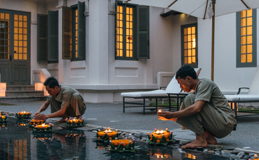Two guests seated at opposite ends of a reflecting pool at Amantaka, with the colonial architecture and lit windows mirrored in the still water.