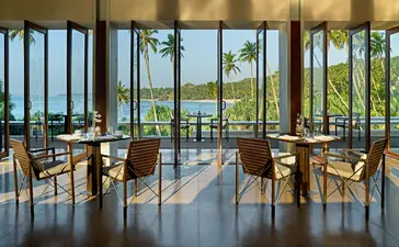 Amanwella dining venue with floor-to-ceiling windows overlooking coastal views and wooden chairs at tables.
