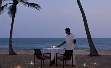 A guest sits at a dining table on Amanwella's beach at dusk, surrounded by candlelit lanterns and swaying palm trees.