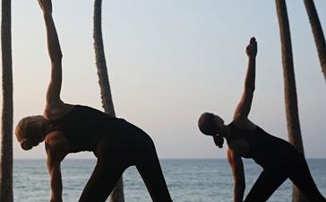 Two figures practising yoga on a wooden deck beneath palm trees overlooking the ocean at Amanwella.