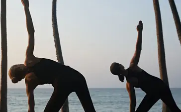 Two figures practising yoga on a wooden deck beneath palm trees overlooking the ocean at Amanwella.