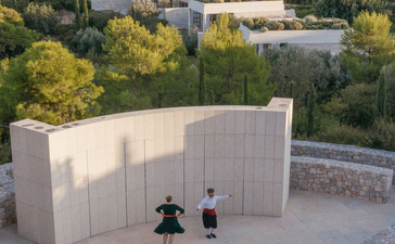 Aerial view of Amanzoe's contemporary pavilion with guests gathered in the courtyard, surrounded by Mediterranean landscape and sea views.