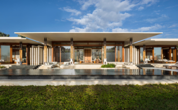 Amanera villa with wooden architecture and wide veranda overlooking manicured gardens under clear sky.