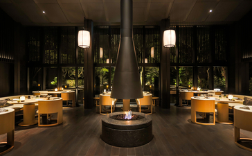 Aman Kyoto's Living Pavilion interior at evening, featuring warm lighting, central fireplace, and symmetrical wooden seating.