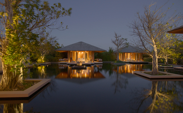 Four-bedroom ocean villa at Amanyara with illuminated pavilions reflected in still water at dusk.