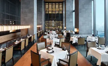 Aman Tokyo's dining venue with wooden flooring, white-dressed tables, and floor-to-ceiling windows overlooking the city.