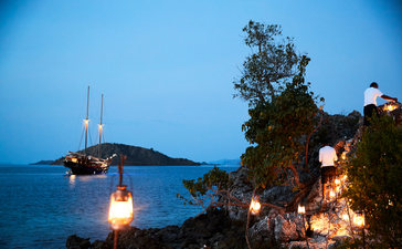 Amandira's waterfront at dusk with illuminated lanterns and a sailboat anchored offshore.