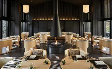 Aman Kyoto resort's Living Pavilion dining space with symmetrical wooden tables, central fireplace, and soft pendant lighting.