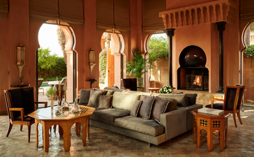 Al Hamra Maison lounge at Amanjena with arched doorways opening to gardens, featuring a grey sofa and wooden furnishings.