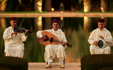 安缦珍纳度假村夜间露台上，三位演奏者坐在椅子上进行传统音乐表演，身后是水池和棕榈树照明。