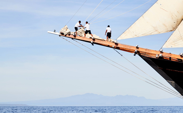 Amandira crew members perched on wooden boat bowsprit extending over calm blue sea.