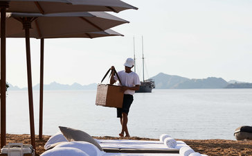 Woman carrying surfboard on beach at Amandira with calm waters and covered seating area in background.