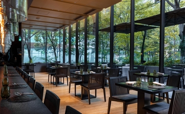 Café at Aman Tokyo with wooden tables, black chairs, and floor-to-ceiling windows overlooking greenery.
