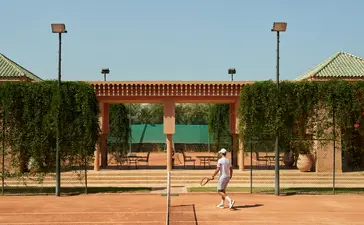 Court de tennis à Amanjena, avec filet et bâtiments aux murs ocre et détails verts.