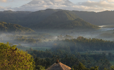 Amankila resort at sunrise, with mist rising over forested valleys and distant mountains under a pale sky.