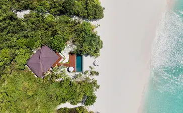 安缦璞乐度假村沙滩池畔别墅的鸟瞰图，展示紫色屋顶建筑与白沙和清澈海水相邻。
