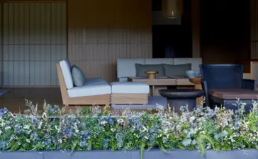 Wooden seating area with white cushions at Amanemu resort, framed by flowering plants.