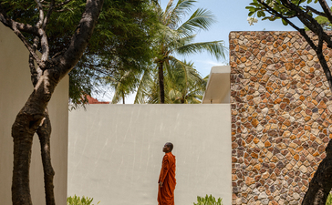 Monk in saffron robes walking through shaded courtyard at Amansara, Cambodia, framed by trees and stone walls.