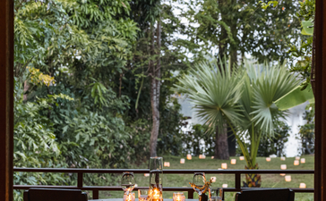 Dining table set for two on a shaded terrace at Amansara, overlooking tropical gardens and palm trees.