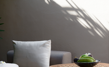 Amanwella suite detail showing cushioned seating, wooden side table with green vessel, and palm shadow on grey wall.