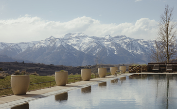 Snow-capped mountains reflected in a still water feature at Amangani, with sparse vegetation and grey sky.