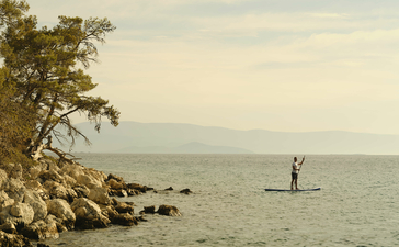 Person paddleboarding on calm waters at Amanruya, Turkey, with rocky shoreline in foreground.
