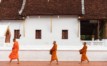 Three Buddhist monks in saffron robes walking past a white temple building at Amantaka.