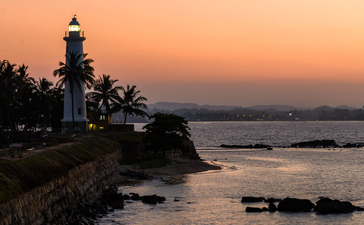Galle Lighthouse stands on a rocky headland at sunset, its beacon illuminated against a soft peachy sky, with palm trees and calm waters below at Amangalla.