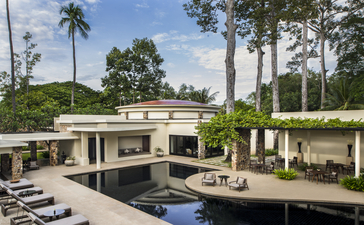 Amansara's main swimming pool surrounded by modernist architecture, manicured gardens, and palm trees under clear blue sky.