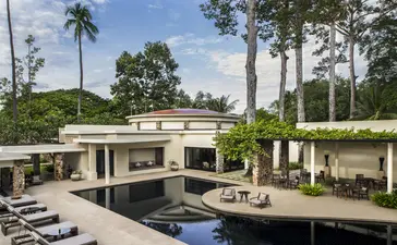 Amansara's main swimming pool surrounded by modernist architecture, manicured gardens, and palm trees under clear blue sky.