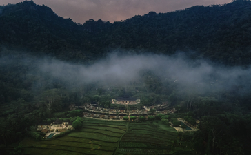 Amanjiwo nestled in a verdant valley at dawn, surrounded by mist-draped mountains under soft, dusky light.