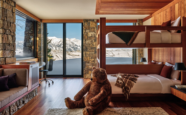 Amangani living room with stone fireplace, wooden shelving, and terrace doors opening to mountain views.