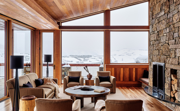 Amangani principal bedroom with expansive windows overlooking snow-capped mountains, featuring wooden beams and stone fireplace.