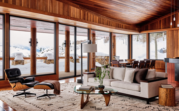 West Tatanka House living room at Amangani with floor-to-ceiling windows overlooking mountain landscape, wood-panelled walls, and mid-century furnishings.