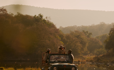 Jeep parked on a riverbank at Aman-i-Khas with forested hills beyond in morning mist.
