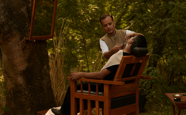 A guest sits alone in a wooden chair beneath towering trees at Aman-i-Khas, surrounded by dappled sunlight filtering through the canopy.