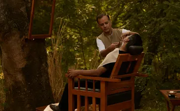 A guest sits alone in a wooden chair beneath towering trees at Aman-i-Khas, surrounded by dappled sunlight filtering through the canopy.